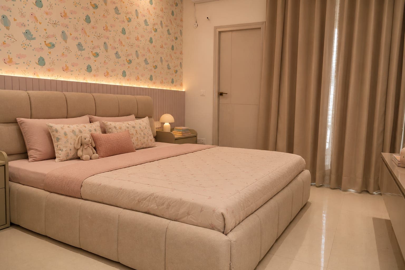 Children's bedroom with pastel bird wallpaper, a cushioned headboard, pink bedding and taupe floor-length curtains.