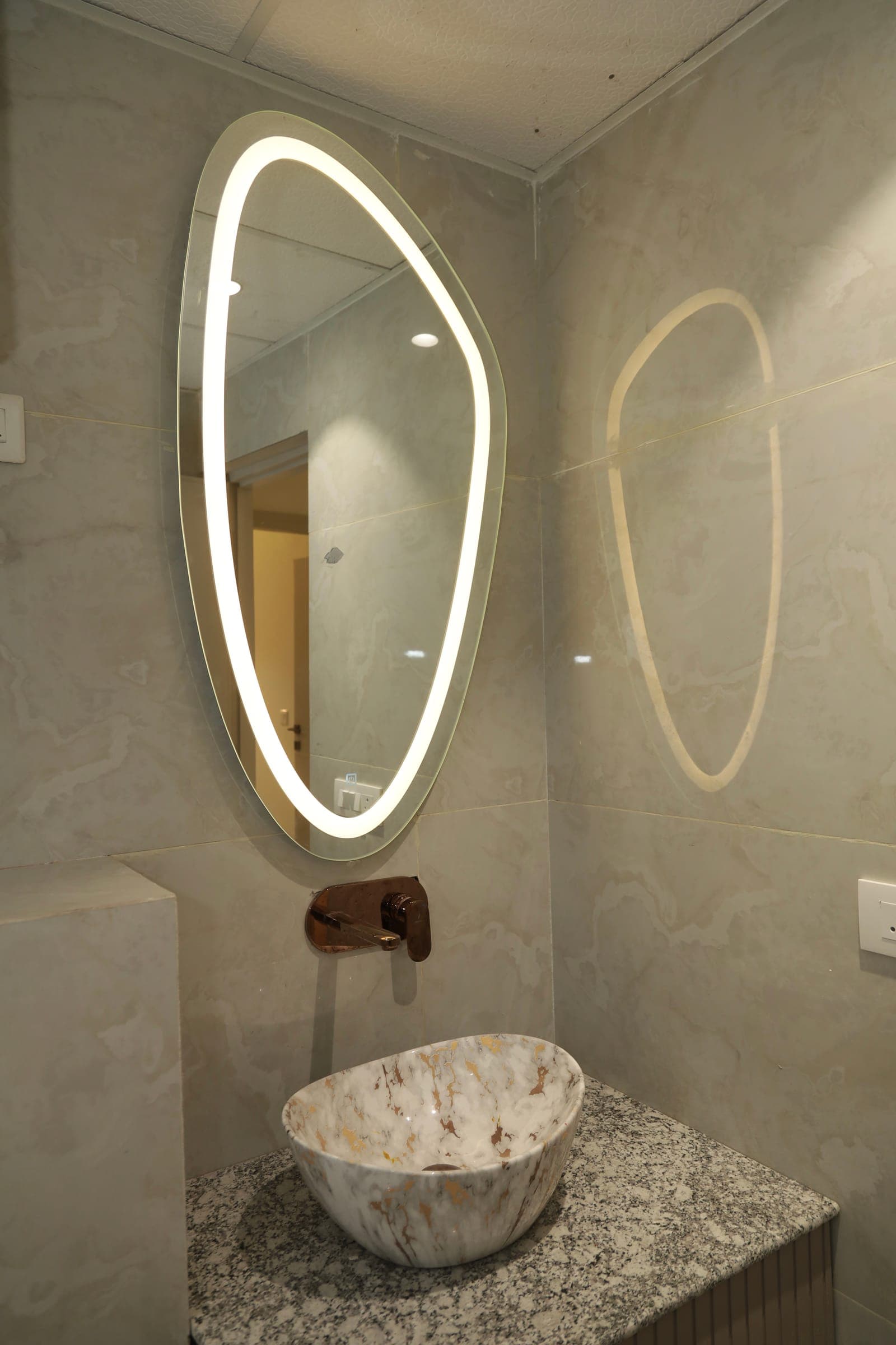 Washroom with an organic-shaped backlit mirror, marble-look walls and a veined stone vessel basin on a stone counter.