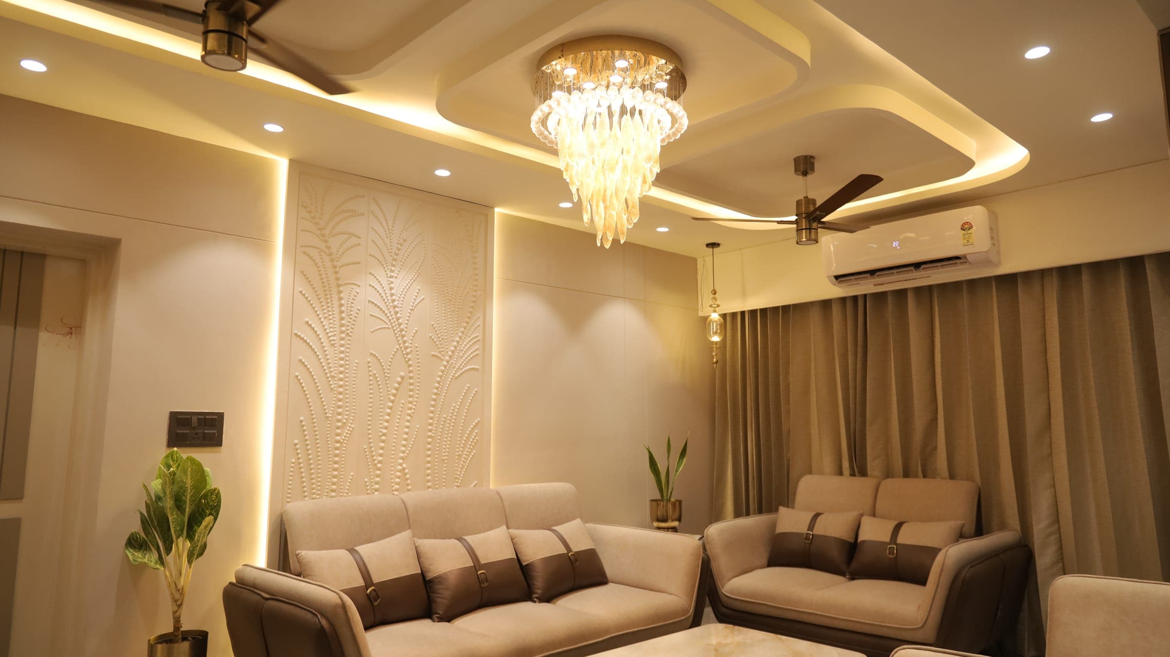Living room seen across two beige sofas, with a sculptural glass chandelier, a cove-lit ceiling, a raised botanical feature panel and floor-length taupe curtains.