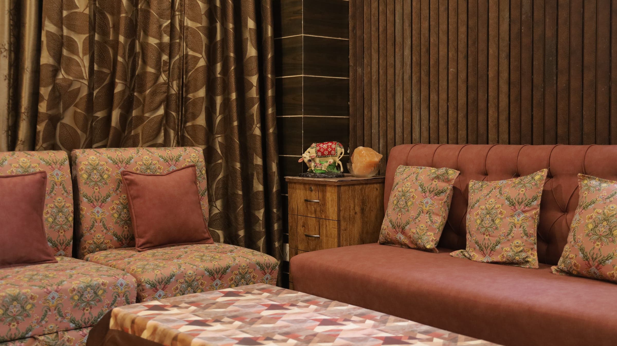 Sitting area with a rust velvet couch and a floral-patterned settee, patterned brown curtains and a fluted wood wall around a geometric-top coffee table.