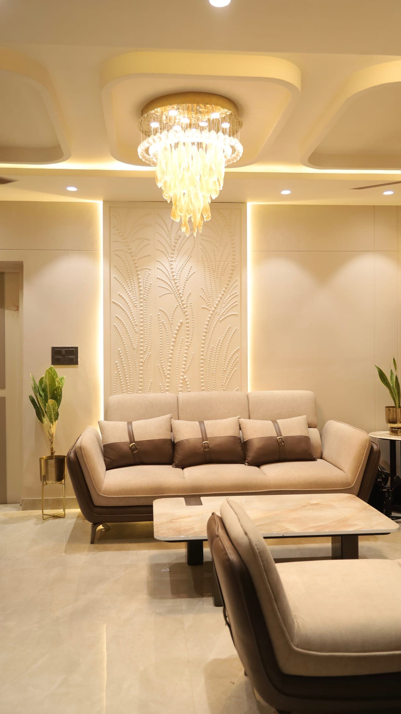 Living room with a three-seat cream sofa below a beaded botanical feature panel, a glass drop chandelier and a marble coffee table on pale stone flooring.
