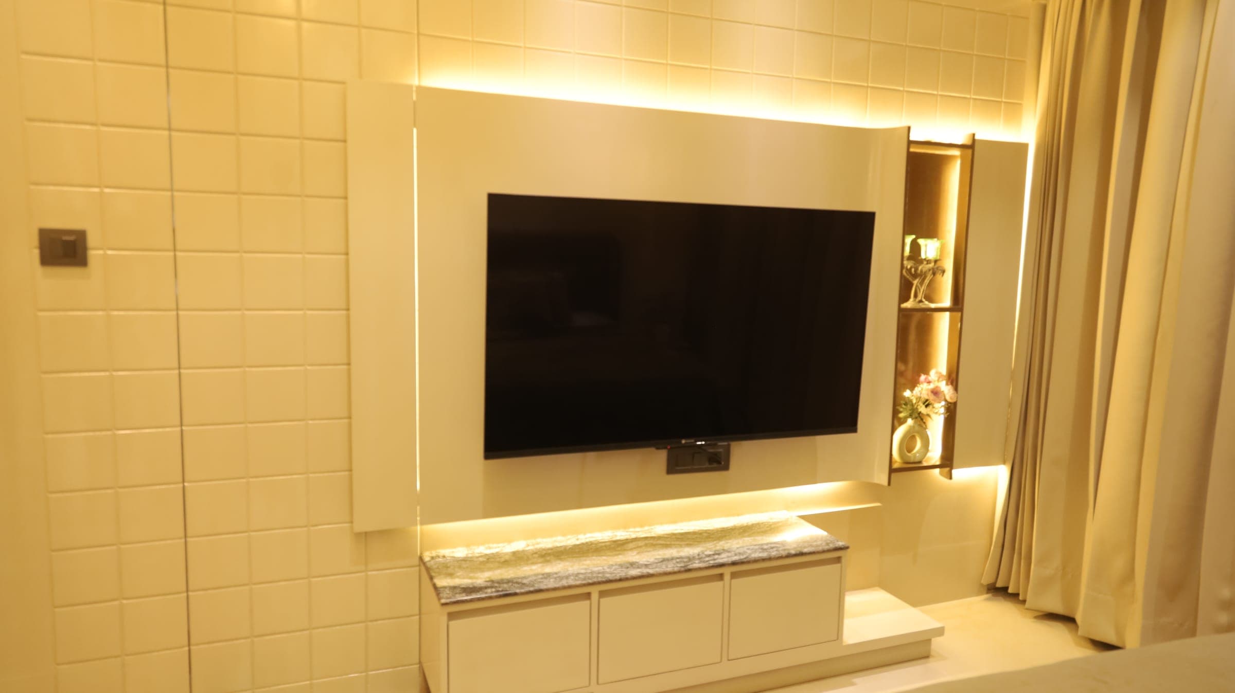 Bedroom television wall in cream tile with a wall-mounted TV, a backlit display niche of decor and a marble-topped cabinet under concealed lighting.