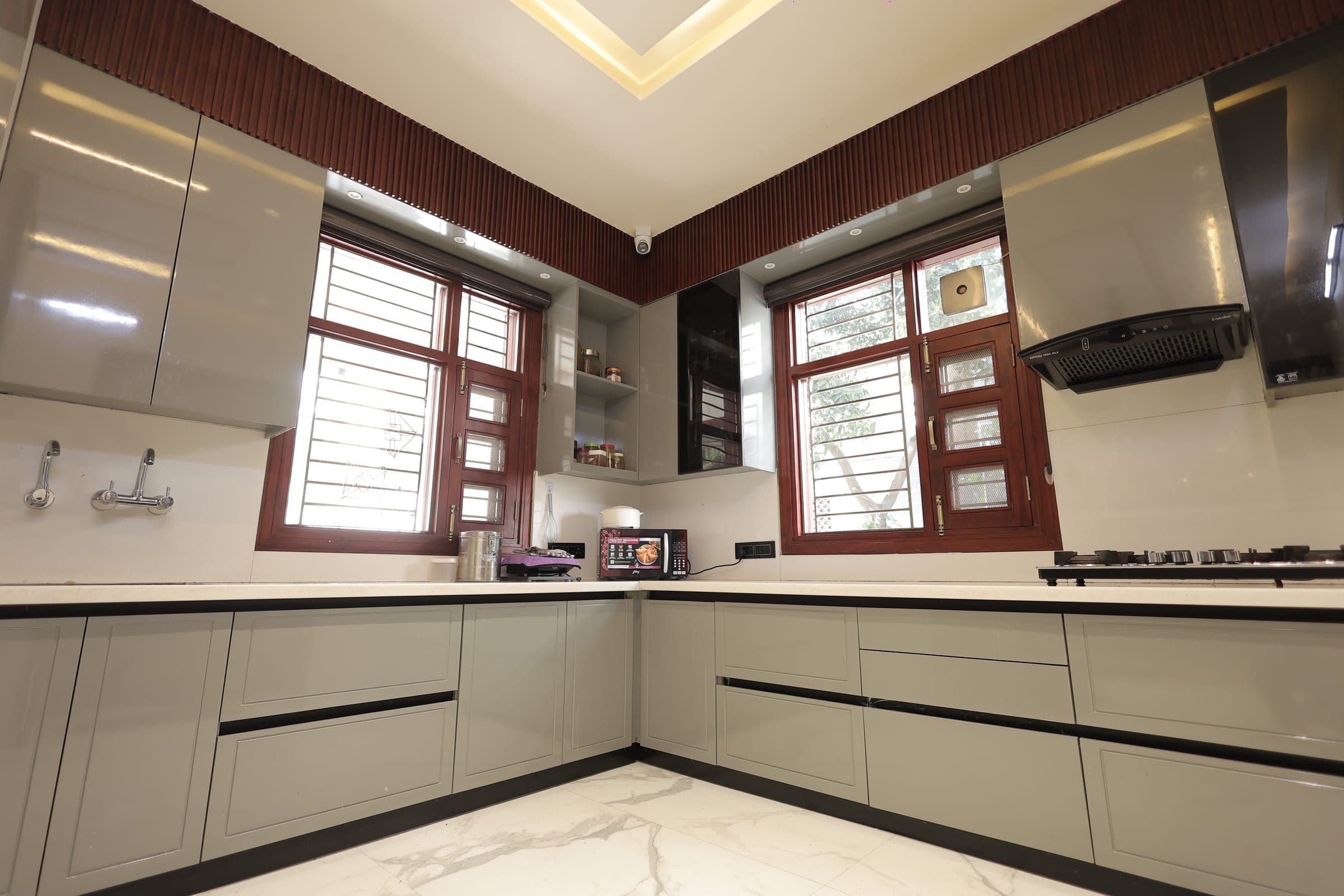 Corner kitchen with grey glossy and shaker cabinets, a marble-look countertop and floor, two wood-framed windows, a black chimney over the hob and warm daylight.