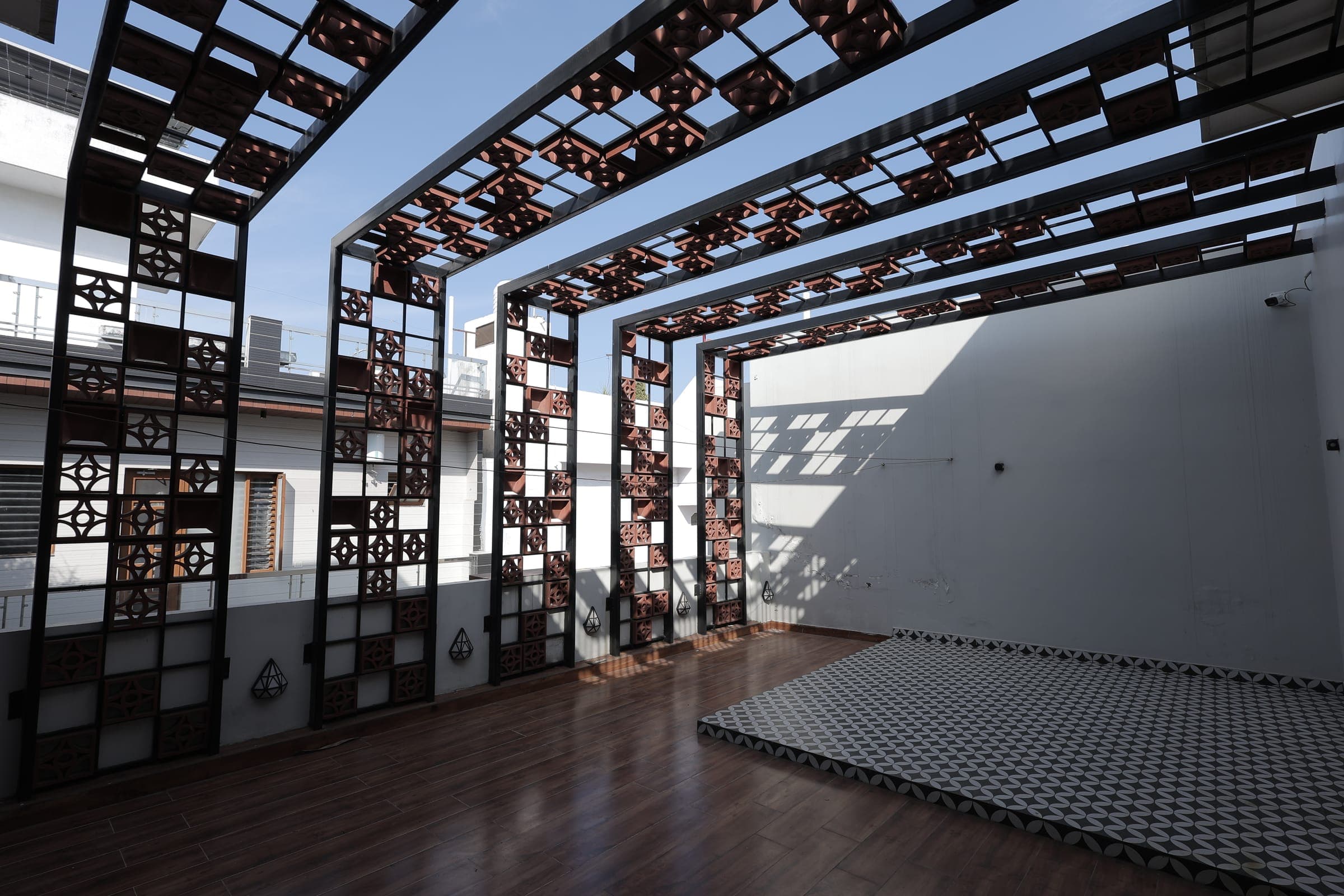 Open terrace with a black steel and terracotta jali pergola overhead, polished timber decking, a patterned black-and-white tiled platform and clear blue sky.