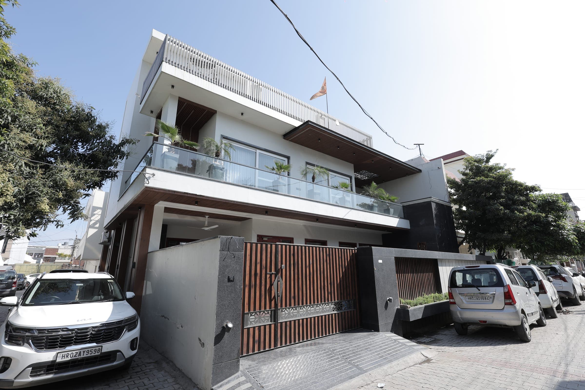 Two-storey house exterior by day with a white and timber facade, glass-railed balconies holding potted palms and cars parked along the street.