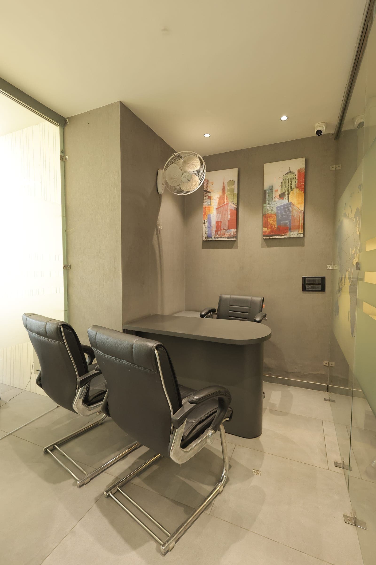 Two black leather visitor chairs facing a desk in a glass-partitioned cabin, with framed cityscape prints and a wall fan on a grey stone wall.