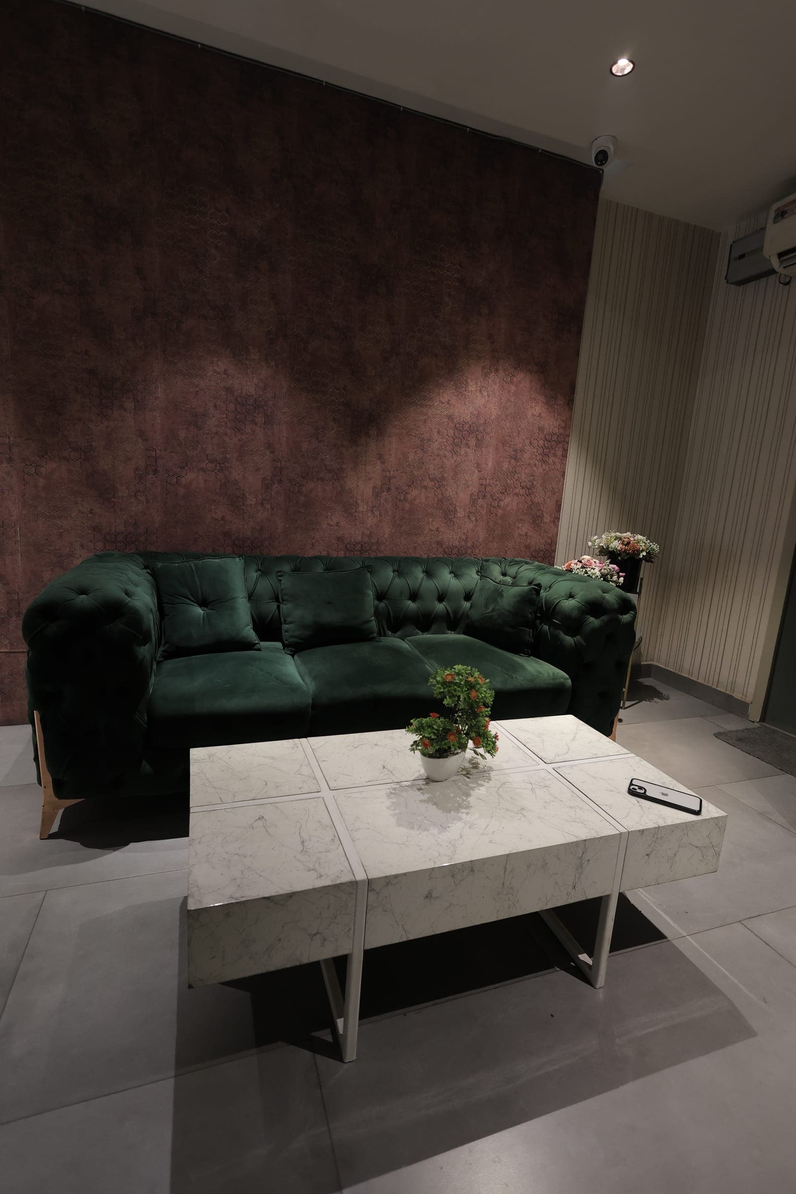 Deep green tufted velvet sofa against a textured maroon feature wall, with a white marble side table and a small vase of orange flowers.